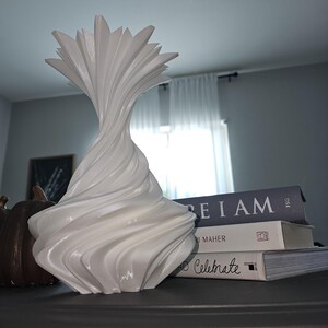 May include: A white, sculptural vase with a unique, swirling design. The vase has a flared top and a textured, organic shape. The vase is displayed on a dark surface, next to a stack of books.
