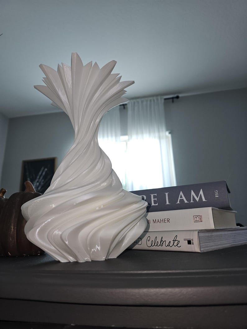 May include: A white, sculptural vase with a unique, swirling design. The vase has a flared top and a textured, organic shape. The vase is displayed on a dark surface, next to a stack of books.