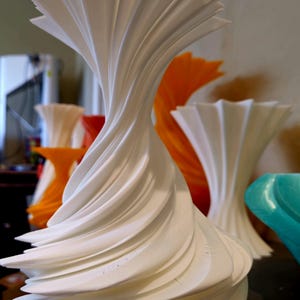 May include: A white, abstract sculpture with a swirling, layered design. The sculpture has a flared top and a rounded base. Other sculptures in orange, white, and teal are visible in the background. The sculptures appear to be made of a solid material.