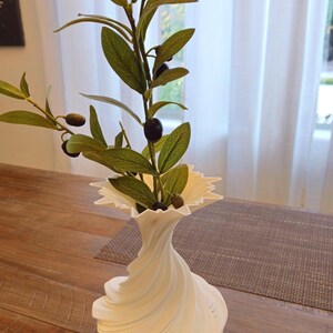 May include: A white, decorative vase with a unique, swirling design, holding olive branches with green leaves and black olives. The vase has a ruffled top and sits on a wooden table. The overall aesthetic is modern and natural.
