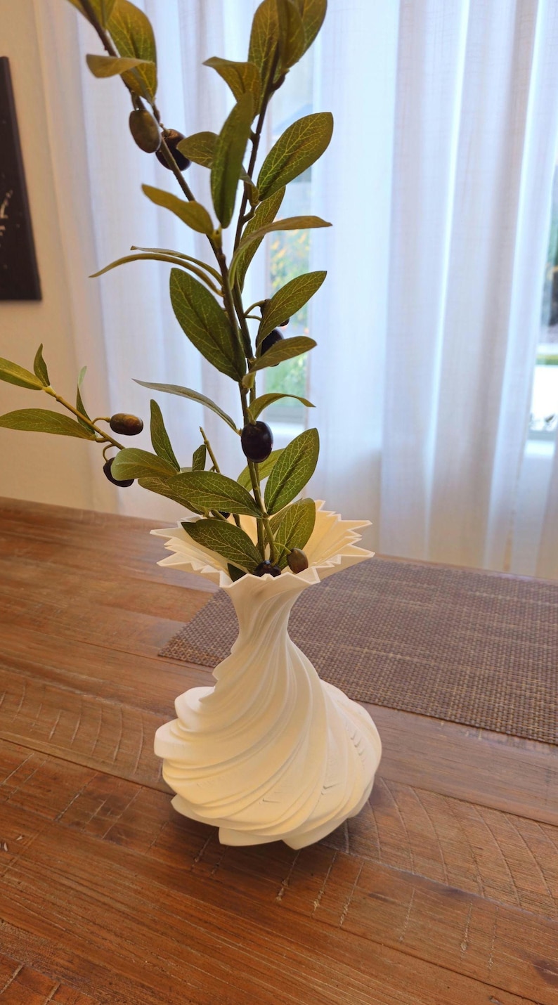 May include: A white, decorative vase with a unique, swirling design, holding olive branches with green leaves and black olives. The vase has a ruffled top and sits on a wooden table. The overall aesthetic is modern and natural.
