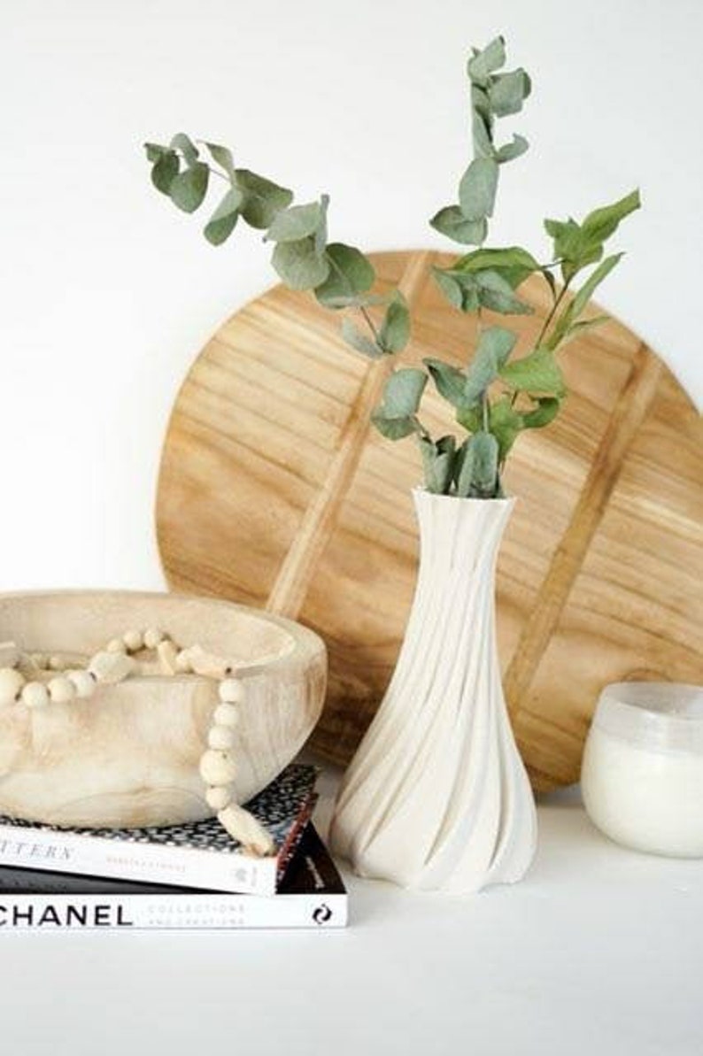 May include: A white, textured vase holds green eucalyptus branches, set against a wooden cutting board and a wooden bowl with beads. A stack of books with the word "CHANEL" is visible.