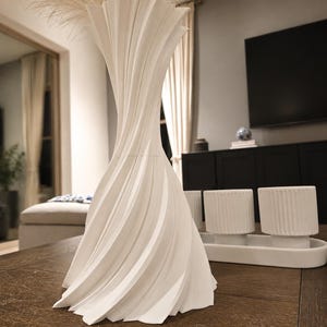 May include: A tall, white, 3D-printed vase with a spiraling, twisted geometric design that tapers toward the center and flares at the top and bottom. The surface features sharp, vertical ridges that create a dynamic, pleated appearance throughout.