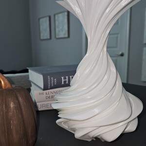 May include: A white, abstract decorative vase with a swirling, layered design. The vase has a flared top and a twisted body. A brown pumpkin and books are in the background.