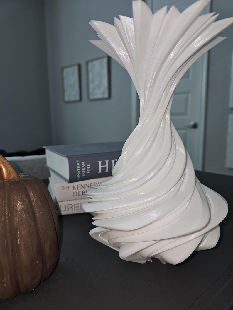 May include: A white, abstract decorative vase with a swirling, layered design. The vase has a flared top and a twisted body. A brown pumpkin and books are in the background.