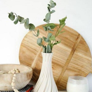 May include: A white, textured vase with a spiral design holds green eucalyptus branches. A wooden bowl with a beaded strand, a wooden cutting board, and a candle in a glass are also present, creating a minimalist, natural aesthetic.