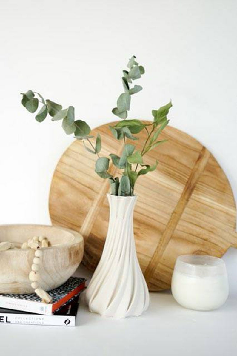 May include: A white, textured vase with a spiral design holds green eucalyptus branches. A wooden bowl with a beaded strand, a wooden cutting board, and a candle in a glass are also present, creating a minimalist, natural aesthetic.