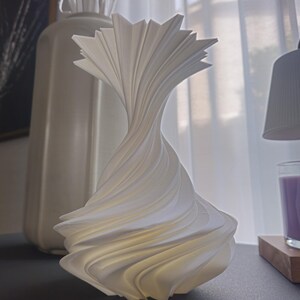 May include: A white, sculptural lamp with a swirling, layered design. The lamp has a unique, abstract shape and is made of a smooth, possibly ceramic material. The lamp is on a dark surface, with a vase and branches in the background.