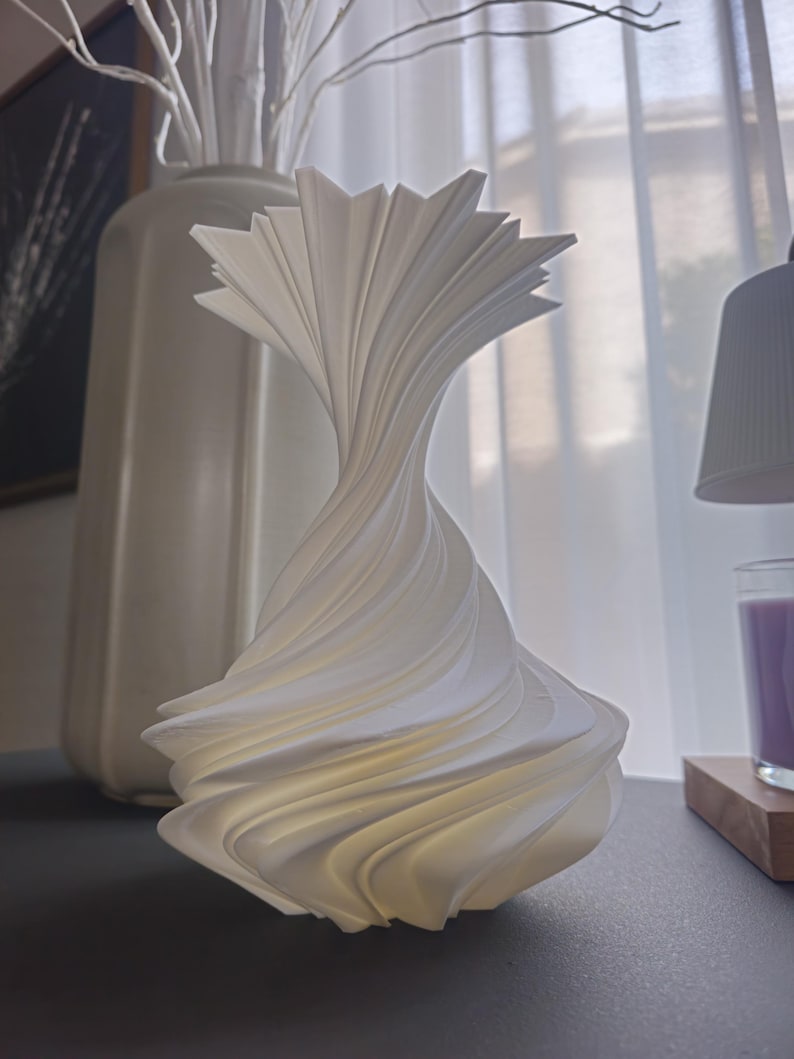 May include: A white, sculptural lamp with a swirling, layered design. The lamp has a unique, abstract shape and is made of a smooth, possibly ceramic material. The lamp is on a dark surface, with a vase and branches in the background.