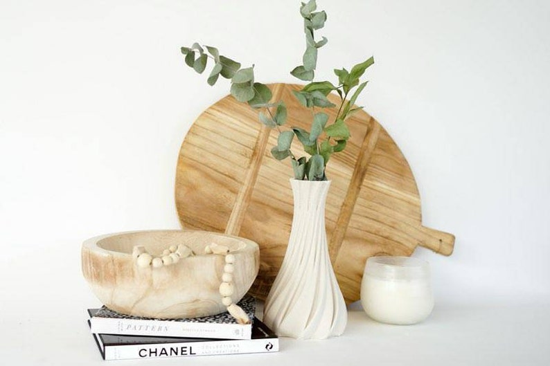 May include: A still life arrangement featuring a wooden cutting board, a white vase with eucalyptus, a wooden bowl with beads, a candle in a glass holder, and two books. The items are arranged on a white surface.