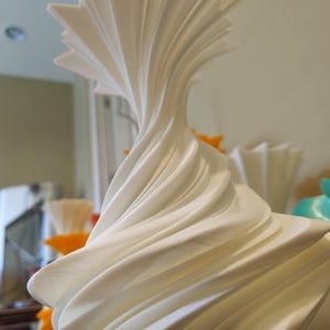 May include: A white, abstract sculpture with a swirling, layered design. The top of the sculpture has a starburst shape. The piece is made of a smooth, solid material and is likely a decorative item.