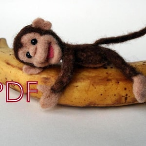 Può includere: Un giocattolo di scimmia di feltro marrone sdraiato su una banana gialla. La scimmia ha un viso sorridente con guance rosa e una bocca rossa. La coda della scimmia è avvolta attorno alla banana.