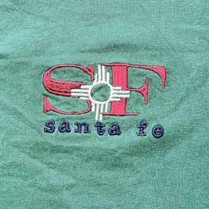 May include: Green fabric with an embroidered design. The design features the letters "SF" in red, with a white and red symbol in the centre, and the words "santa fe" embroidered in blue below.