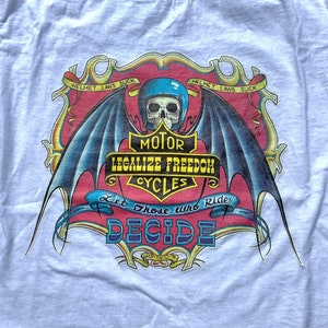 May include: A white t-shirt featuring a graphic print of a skull with wings and a banner that reads "Motor Legalize Freedom Cycles Let Those Who Ride Decide". The banner is red and gold with black lettering. The skull is wearing a blue helmet. The graphic is enclosed within a gold frame with the words "Helmet Laws Suck" at the top and bottom.