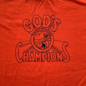 May include: Orange t-shirt with a blue graphic design that says "God's Champions" with a shield and sword design that says "Equipped to Follow Jesus".