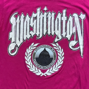 May include: A pink t-shirt with a white graphic of the word "Washington" in a gothic font. The graphic also includes a white circle with the words "Washington D.C. Capital of the United States" and a white wreath.