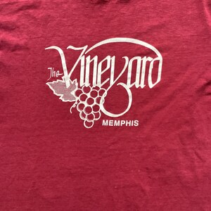 May include: A maroon t-shirt with a white graphic of a stylized grapevine with the text "The Vineyard Memphis".