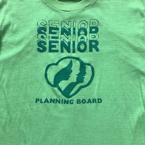 May include: Green t-shirt with the word "SENIOR" repeated four times in a stacked design. The shirt also features a green Girl Scout logo with the words "PLANNING BOARD" below it.