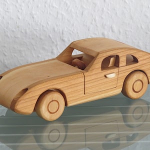May include: A wooden toy car with a light brown finish. The car has a rounded body, four wheels, and a detailed interior.