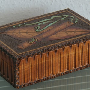 May include: A wooden cigar box with a carved design of a cigar and a tobacco leaf. The box has a hinged lid and a decorative border.