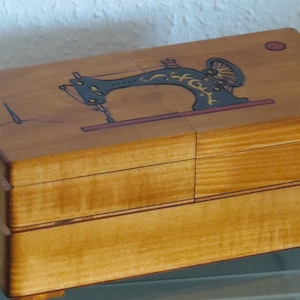 Op de afbeelding: Een houten doos met een scharnierend deksel. Het deksel heeft een geschilderd beeld van een vintage naaimachine met gouden accenten. De doos heeft een natuurlijke houtkleur en twee kleine rode cirkels op de bovenkant.