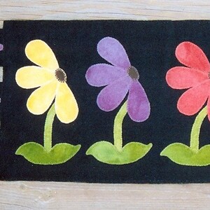 May include: Black rectangular table runner with colorful flower appliqués. The flowers are yellow, purple, and red with green stems and leaves. The runner has decorative purple teardrop shapes on each end.