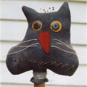 Może przedstawiać: Czarna filcowa poduszka na szpilki w kształcie kota z czerwonym filcowym nosem, żółtymi guzikowymi oczami i czarnymi szwami wokół ust. Poduszka na szpilki jest przymocowana do drewnianej szpulki.