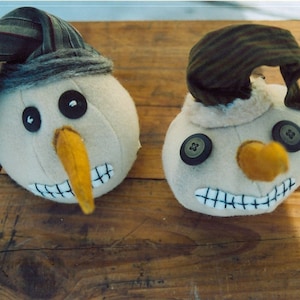 May include: Two whimsical snowman ornaments. Each has a beige head with black button eyes, a yellow carrot nose, and a stitched mouth. They wear hats made from patterned fabric. One hat is green and gray striped, the other is brown and green striped.