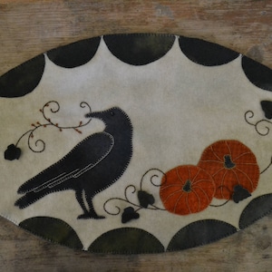 May include: A black crow with a long beak sits on a white felt background with a black and orange pumpkin design. The crow is embroidered with black thread and the pumpkins are embroidered with orange thread.