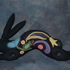 May include: Black felt rabbit with colorful embroidered details, including flowers, leaves, and swirls. The rabbit is in a running pose with its ears perked up.