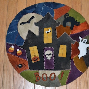 Op de afbeelding: Een ronde, veelkleurige wandhanger van vilt met een gestikt ontwerp van een spookhuis, een zwarte kat, een geest, snoepmais, een spin en het woord "BOO!"