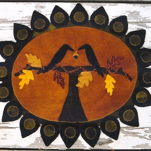 May include: A felt wall hanging with a brown oval center featuring two black birds perched on a tree branch with yellow and brown leaves. The oval is surrounded by a black and green sunburst design.
