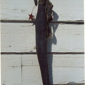 May include: A black fabric doll with a long, thin body and a star-shaped decoration. The doll is hanging on a white wooden wall.