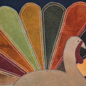 Op de afbeelding: Een bruine en beige vilt kalkoen met een donkerblauwe achtergrond. De kalkoen heeft een grote waaier van veren in tinten bruin, groen en oranje.