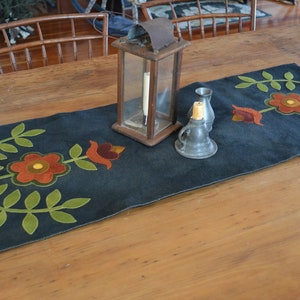 May include: A dark blue table runner with a floral design featuring green leaves and red, orange, and yellow flowers. The runner is on a wooden table with a lantern and a small oil lamp.