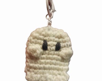 Puede incluir: Un llavero de fantasma tejido a ganchillo en color blanco, con dos pequeños ojos bordados en negro y dos brazos redondeados a los lados. El fantasma está unido a un mosquetón de plástico blanco mediante un cordón corto de color blanco.
