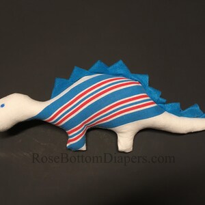 May include: A white plush dinosaur toy with blue and red striped accents on its back and body. The dinosaur has a blue felt spine and a small blue eye. The website RoseBottomDiapers.com is printed on the bottom.