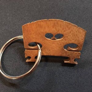May include: A wooden violin bridge keychain with a silver-colored key ring. The bridge is a light brown color and has a curved top with two oval-shaped cutouts. The keychain is likely a music-themed accessory.