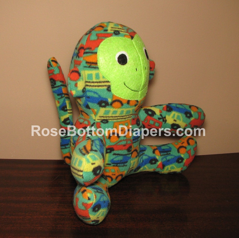 May include: A plush monkey toy with a green face and a body covered in a colorful pattern of cars, trucks, and buses. The toy has black eyes and a stitched smile. The text "RoseBottomDiapers.com" is visible on the bottom.
