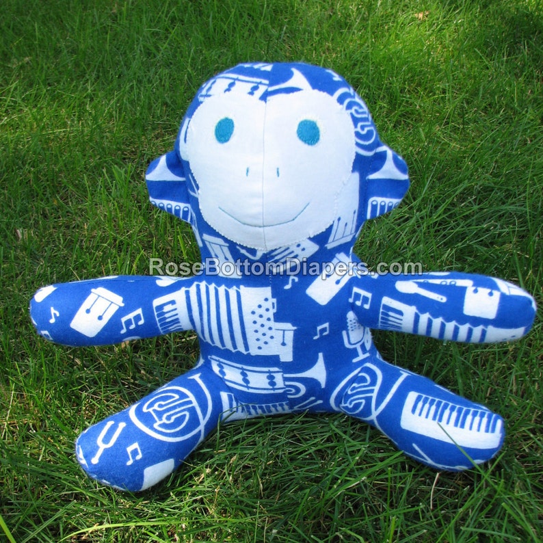 May include: A blue plush monkey with white musical instrument patterns. The monkey has a white face, blue eyes, and is lying on green grass. The text "RoseBottomDiapers.com" is visible at the bottom of the image.
