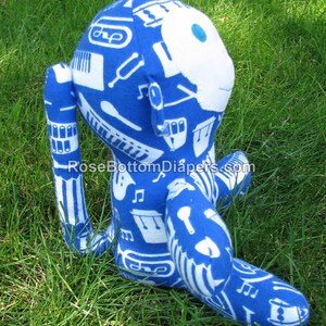 May include: A blue plush monkey toy with white musical instrument patterns. The monkey has a white face with a blue dot for an eye. The toy is sitting on green grass.