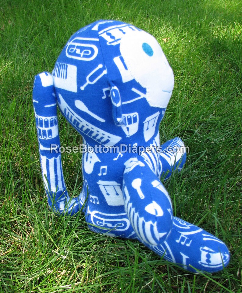 May include: A blue plush monkey toy with white musical instrument patterns. The monkey has a white face with a blue dot for an eye. The toy is sitting on green grass.