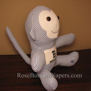 May include: A stuffed monkey doll with blue and white striped fabric. The monkey has a white face, brown eyes, and a small smile. A patch on the chest displays the letter "B" and the word "Berkeley". The doll is sitting with its arms and legs extended.
