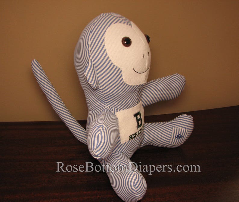 May include: A stuffed monkey doll with blue and white striped fabric. The monkey has a white face, brown eyes, and a small smile. A patch on the chest displays the letter "B" and the word "Berkeley". The doll is sitting with its arms and legs extended.