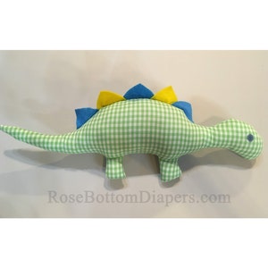 May include: A stuffed dinosaur toy in a green and white gingham pattern. The dinosaur has blue and yellow felt spikes along its back and a small blue eye. The toy is designed for children.