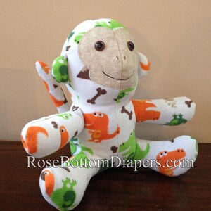 May include: A handmade plush monkey with a white background and a pattern of orange and green dinosaurs, bones, and frogs. The monkey has a tan face, brown button eyes, and is sitting on a wooden surface. The text "RoseBottomDiapers.com" is visible.