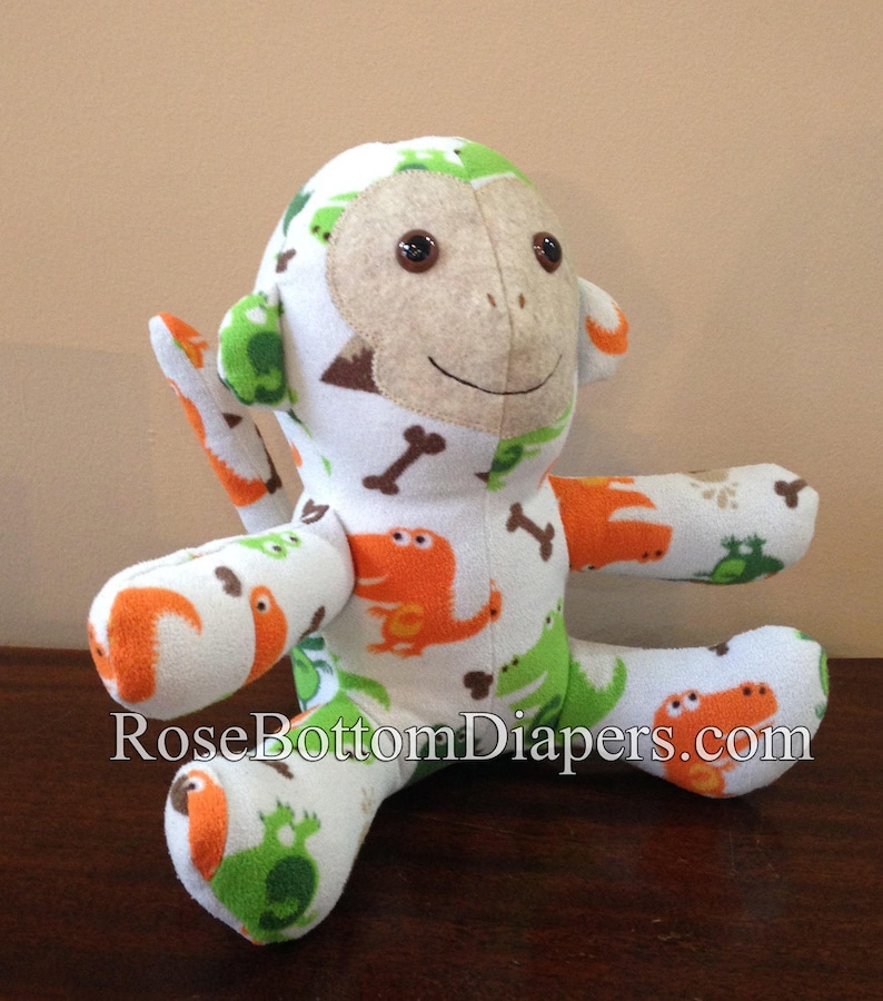 May include: A handmade plush monkey with a white background and a pattern of orange and green dinosaurs, bones, and frogs. The monkey has a tan face, brown button eyes, and is sitting on a wooden surface. The text "RoseBottomDiapers.com" is visible.