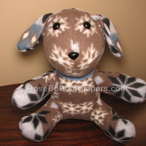 May include: A soft toy dog with a brown body, snowflake pattern, and black eyes. The dog has blue accents on its ears and a blue collar. The toy is on a wooden surface.