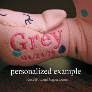 May include: A pink plush toy with the name "Grey" embroidered in pink, along with the date "04/24/14". The toy has blue and teal polka dots and a small embroidered face. The text "personalized example" is at the bottom.
