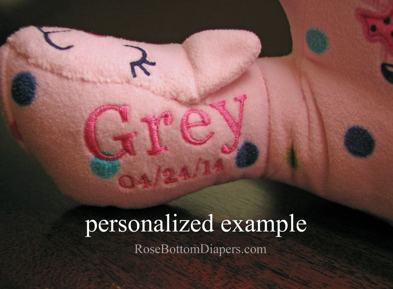 May include: A pink plush toy with the name "Grey" embroidered in pink, along with the date "04/24/14". The toy has blue and teal polka dots and a small embroidered face. The text "personalized example" is at the bottom.